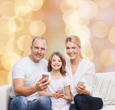 family, holidays, technology and people concept - smiling mother, father and little girl with smartphones over beige lights background