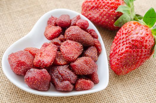 Dried strawberries in heart shape bowl with fresh strawberries on sack background.
