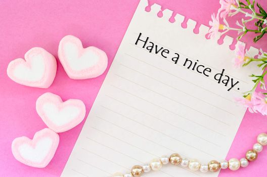 Have a nice day written with sweet heart shape of pink marshmallows with flower on pink background.