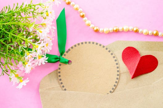 Blank brown paper tag and red heart paper with flower on pink background.