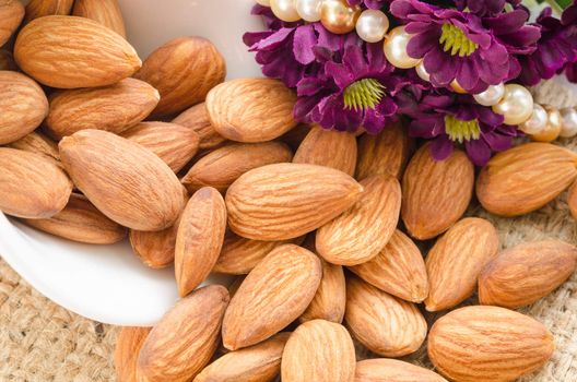 Raw almonds spilling out of small white bowl with flower on sack background.