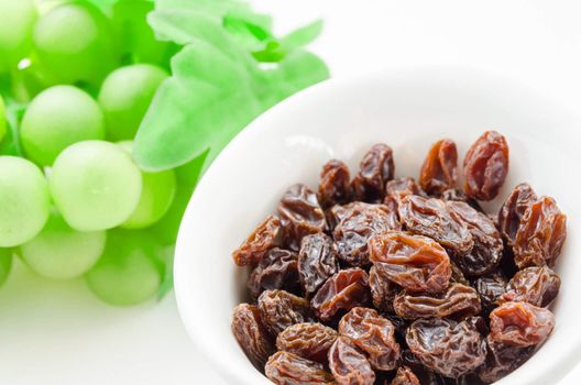 Raisins in white bowl and green grapes on white background.