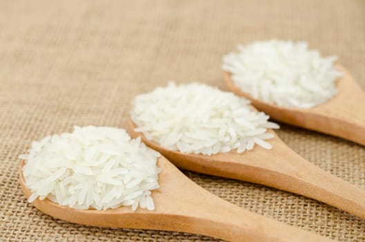 White uncooked rice in small wooden spoon on sack background.