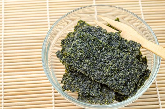 Fried seaweed in glass cup with wood fork on bamboo mat background.