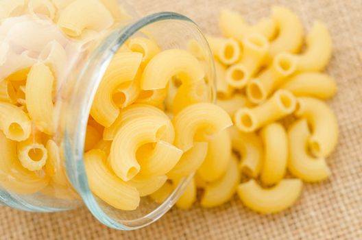 Elbow macaroni noodles spilling out of a glass on sack background.