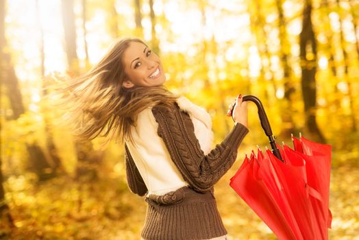 Beautiful cheerful young woman with red umbrella having fun in sunny forest in autumn colors.