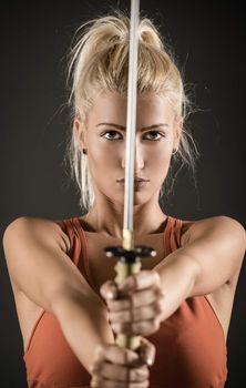 Pretty blonde girl with a serious expression on her face holding a katana outstretched hands and looking at the camera. Selective focus.