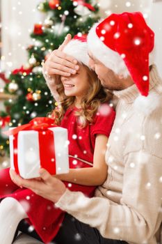 christmas, holidays, family and people concept - smiling father and daughter in santa hats holding gift box and covering eyes at home