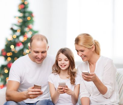 family, holidays, technology and people - smiling mother, father and little girl with smartphones over living room and christmas tree background