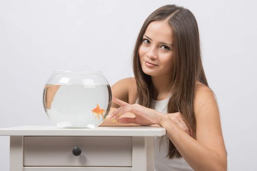 A young girl sits next to a round aquarium in which swimming goldfish