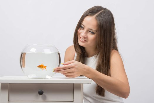 A young girl sits next to a round aquarium in which swimming goldfish