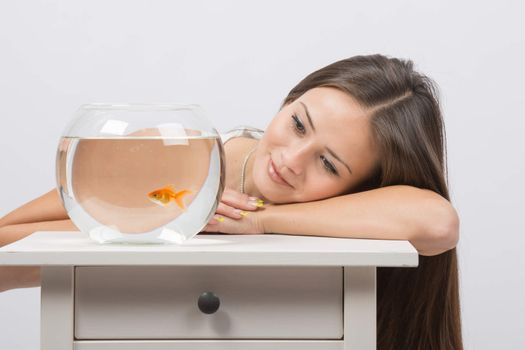 A young girl sits next to a round aquarium in which swimming goldfish