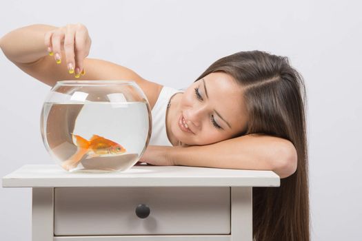 A young girl sits next to a round aquarium in which swimming goldfish