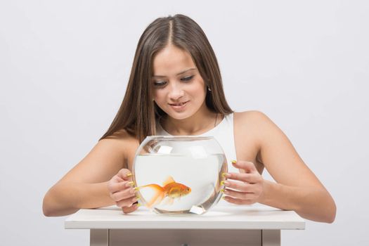 A young girl sits next to a round aquarium in which swimming goldfish