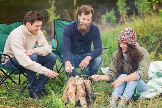 adventure, travel, tourism and people concept - group of smiling friends with marshmallow sitting around bonfire in camping