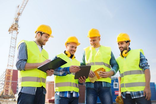 business, building, teamwork, technology and people concept - group of smiling builders in hardhats with tablet pc computer and clipboard outdoors