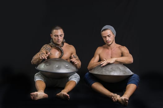 Two Exotic Male Drummers Drumming with Hands on Steel Pan Drums While Seated Beside Each Other in Studio, Isolated on Black Background