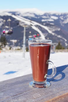 Glass of hot chocolate on wooden table over winter ski resort landscape