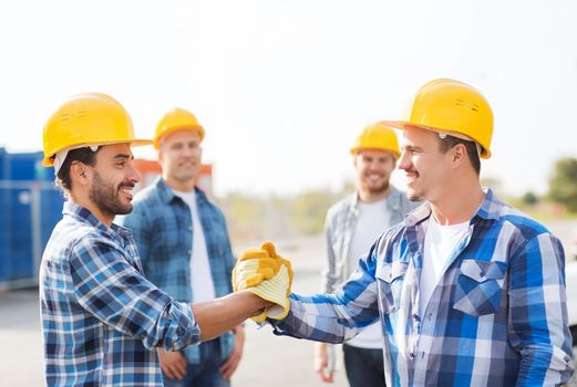 business, building, teamwork, gesture and people concept - group of smiling builders in hardhats greeting each other with handshake outdoors