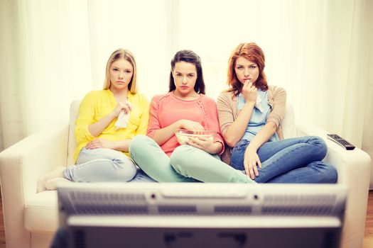 home, technology and friendship concept - three sad teenage girl watching tv at home and eating popcorn