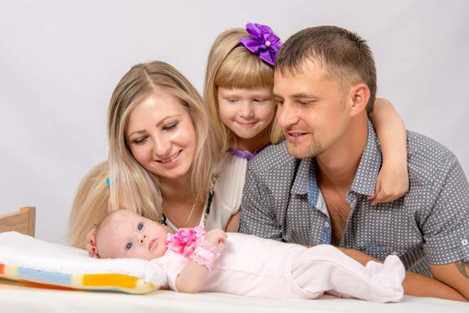 Young parents and daughter sit at the five-year crib, the crib is on a two-month baby on her back