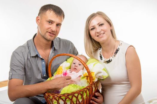 Young parents mom and dad sit on the bed and keep in the hands of his two-month newborn baby girl in a basket