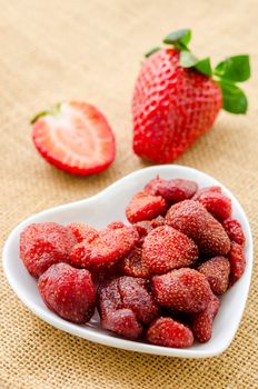 Dried strawberries in white bowl and fresh strawberry on sack background.