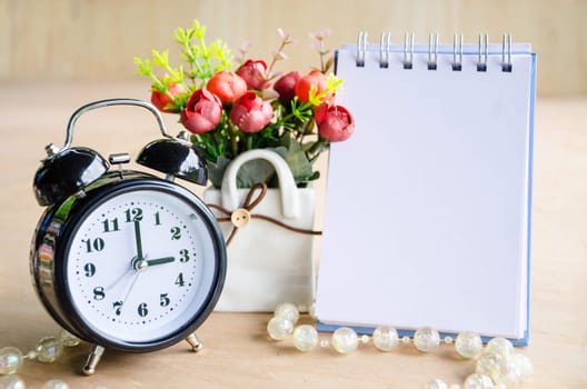 Black alarm clock and open diary with flower on wooden background.