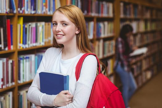 Smiling student with backpack holding a book in library at the university