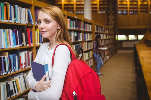 Smiling student with backpack holding a book in library at the university