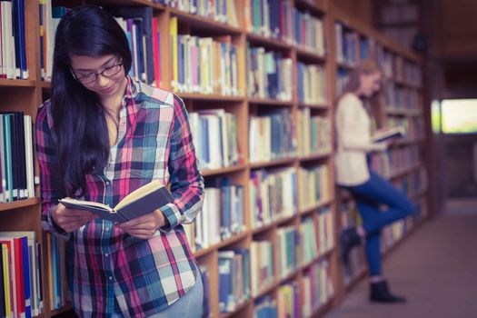 Smiling student leaning against bookshelves reading book at the university