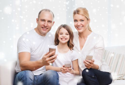 family, home, technology and people concept - smiling mother, father and little girl with smartphones over snowflakes background