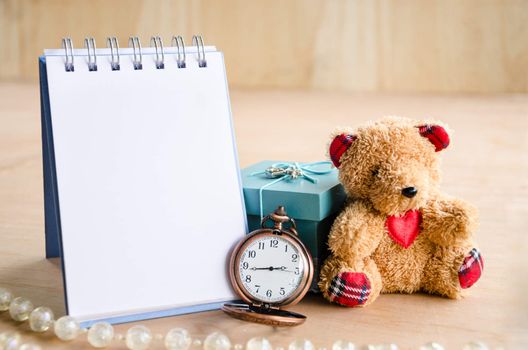 Open diary and vintage pocket watch with brown bear doll on wooden background.