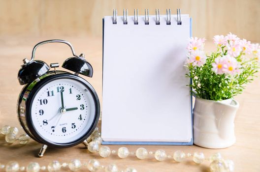 Open diary and alarm clock with flower on wooden background.