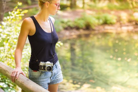 Beautiful blonde caucasian girl wearing jeans shorts an sporty black sleeveless t-shirt, outdoors in nature, carrying vintage camera over her shoulder. Healthy active lifestyle. Square composition.