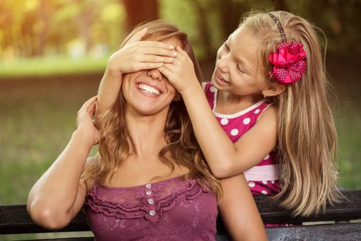 Happy cute little girl and her mother playing in the park. Daughter wants surprise his mom. She is huging mother and holding hands on her eyes.