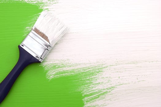 Paintbrush with white paint painting over green colour on a pine board
