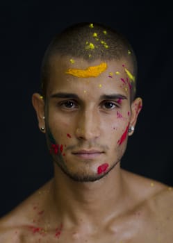 Head and shoulders shot of attractive young man shirtless, skin painted all over with bright Holi colors, looking at camera, isolated on black background