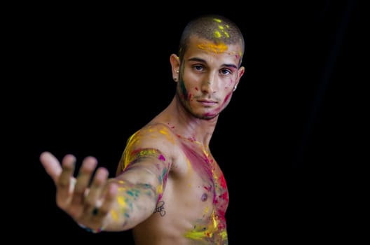 Handsome young man with skin all painted with colors, arm stretched towards camera on dark background