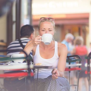 Calm casual blond lady enjoying cup of coffee outdoor in typical italian  street coffee house on warm summer day.
