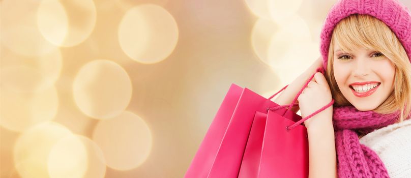 happiness, winter holidays, christmas and people concept - smiling young woman in hat and scarf with pink shopping bags over beige lights background