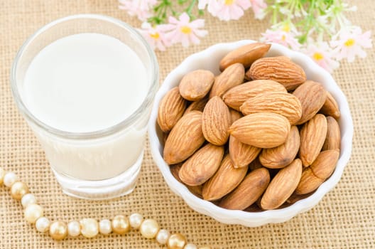 Almond milk in glass and almonds in white cup with flower on sack background.