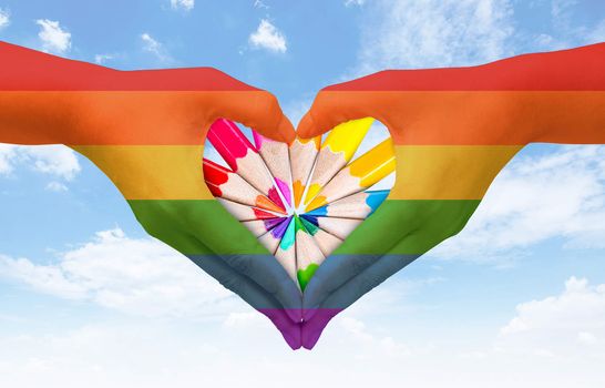 Hands painted as the rainbow flag forming a heart, symbolizing gay love on blue sky. Fredom concept.
