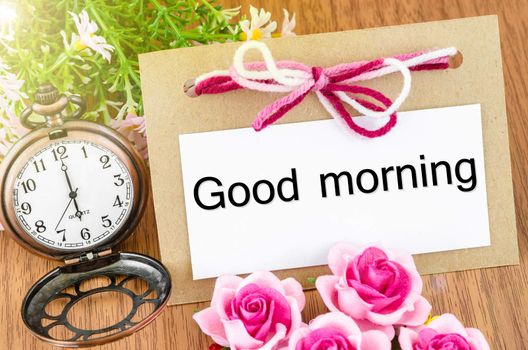 Good morning paper tag and pocket watch with pink rose on wooden background.