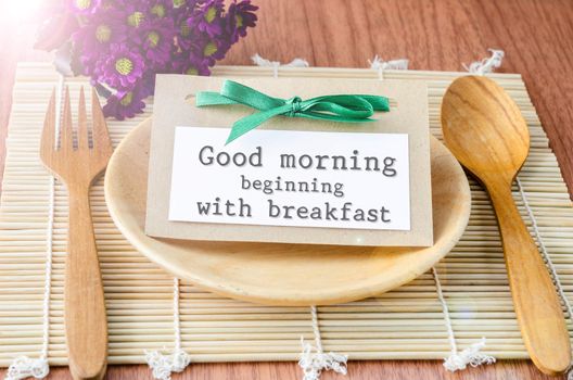 Good morning beginning with breakfast tag on dish spoon with flower on wooden background.