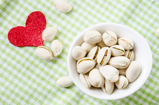 Pistachio nuts in white bowl and red heart shape paper on checkered cloth