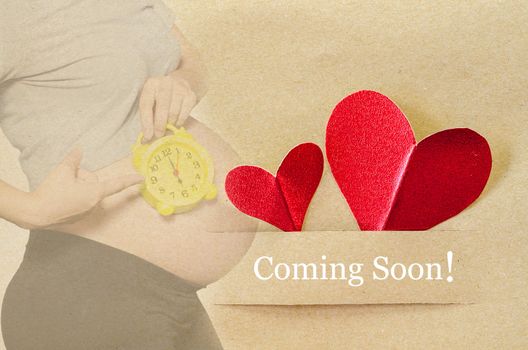 Coming soon. Pregnant woman with red heart on brown paper tag.