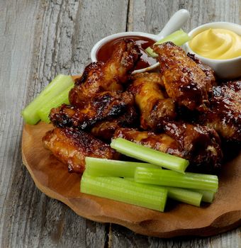 Delicious Chicken Legs and Wings Barbecue with Ketchup and Cheese Sauces and Celery Sticks on Wooden Plate closeup on Rustic Wooden background