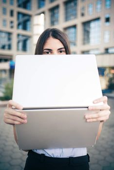 Young beautiful business woman standing in front of office building and hiding behind laptop in her hands.