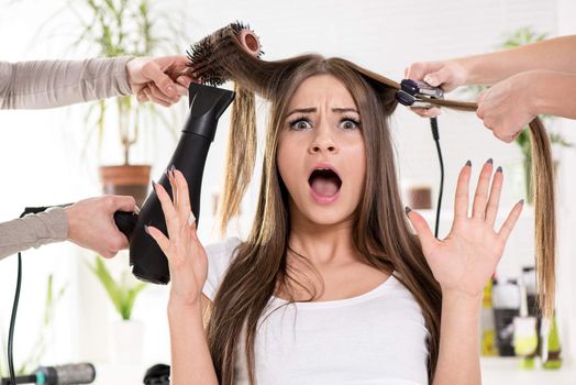 Shocked woman straightening long brown hair with hair dryer and round brush and hair irons at the hairdresser.